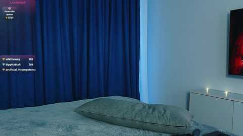 Video of a modern, dimly lit bedroom with a white bed covered in a plush gray blanket. Dark blue curtains cover the left wall, and a white nightstand with a lit candle sits on the right.
