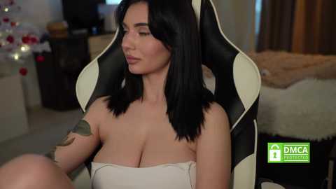 A video shows a curvy woman with black hair, large breasts, and a tattoo on her arm, seated in a black-and-white gaming chair, with a blurred background featuring a Christmas tree and a bed.