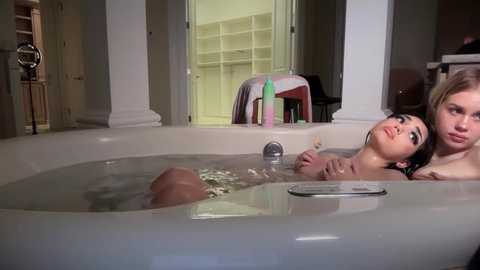 A video of two nude women, one with blonde hair, lying in a bathtub, in a modern bathroom with white walls and glass doors.
