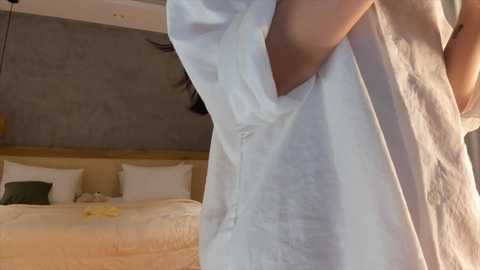 Video of a person in a white robe standing in a modern, minimalist bedroom with a beige bed, green pillows, and gray wall.
