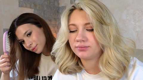 Video of two young women with fair skin, one with straight dark hair, the other with wavy blonde hair. The blonde is brushing her hair while the brunette looks on.