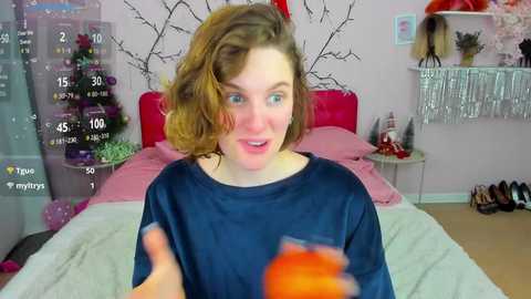 Video of a young woman with wavy brown hair, blue eyes, and a fair complexion, wearing a navy-blue t-shirt, in a pink-themed bedroom with a bed, shelves, and a tree-patterned wall.