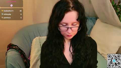A video of a young woman with long black hair, wearing glasses, sitting on a light blue couch with a floral pillow, reading a book.