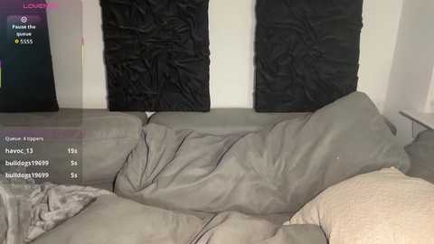Video of a messy, unmade bed with gray sheets and a beige blanket, against a white wall adorned with two black, wrinkled pillows.