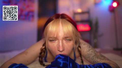 Video of a young woman with blonde and brown hair, freckles, and tattoos, wearing a blue satin dress, performing oral sex on a man. Background shows a blurred room with a colorful poster and red light.