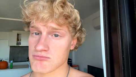 Video of a young, fair-skinned, curly-haired man with a pouty expression, taken indoors near a kitchen.