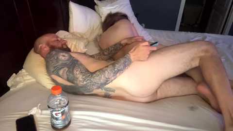 Video of a tattooed, nude man lying on a white bed with a bottle of water, holding a smartphone, in a dimly lit room.