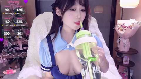 Video of an Asian woman with fair skin, black hair, and red lipstick, wearing a blue maid outfit with suspenders, drinking from a green and white sippy cup, in a cozy, softly-lit room.