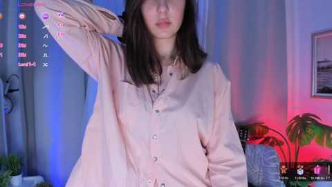 Video of an Asian woman with shoulder-length dark hair, wearing a light pink button-down shirt, in a dimly lit room with a pink and blue light, potted plants, and framed pictures on the wall.