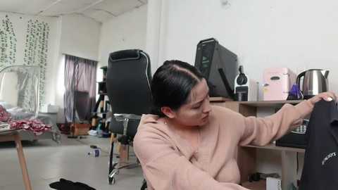 Video of an Asian woman in a pink sweater, sitting in a cluttered, dimly lit room with a black chair, desk, computer, and various household items scattered around.