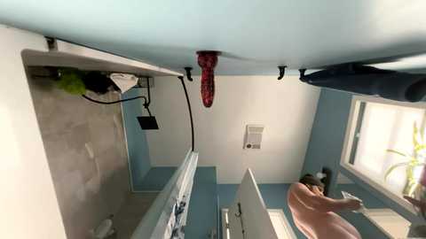 Video of a modern, bright kitchen with light blue walls and white cabinets. A red, textured grip hangs from the ceiling. A person, bare-chested, is reflected in the mirror, wearing dark pants.