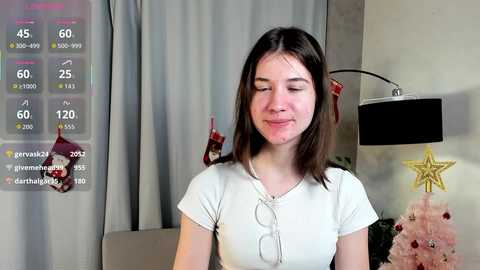 Video of a young woman with fair skin, straight brown hair, and a light complexion, wearing a white t-shirt, smiling in a modern living room with a Christmas tree and red stockings.
