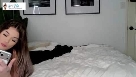 A video of a young woman with long brown hair, wearing a black sweater, lying on a white bed, taking a selfie with a silver phone, in a minimalist, white-walled bedroom with framed black-and-white photos on the wall.
