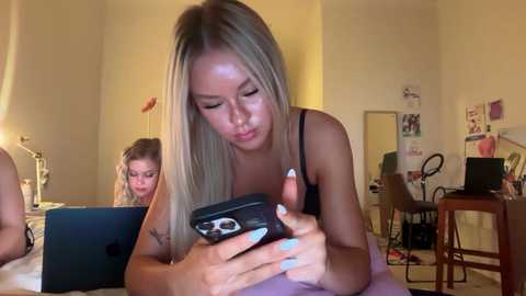 A video captures a young, light-skinned woman with long, straight blonde hair in a black top intently using a smartphone in a cozy, cluttered bedroom.