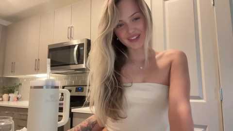 Video of a smiling, slender blonde woman in a cream-colored strapless dress, holding a large white mug, in a modern, beige-toned kitchen with stainless steel appliances and white cabinets.