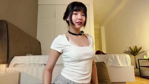 Video of a young Asian woman with short black hair, wearing a white top and gray skirt, standing in a minimalist, modern bedroom with beige walls and a bed.