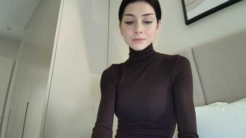 Video of a woman with fair skin, dark hair, wearing a black turtleneck sweater, standing in a modern bedroom with light-colored walls and a beige upholstered headboard.
