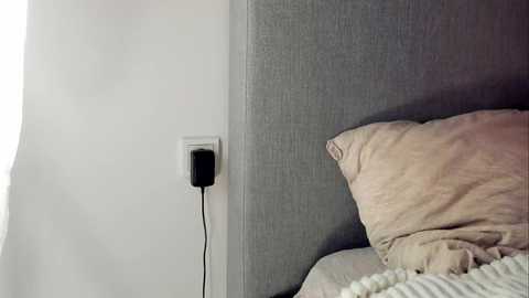 Video of a minimalist bedroom corner featuring a gray upholstered headboard, beige pillows, and a white wall. A black electrical outlet and a charging cable are visible on the left wall.