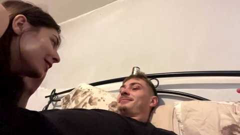 A video shows a young man with short brown hair wearing a black shirt lying on a bed with beige pillows, smiling at a woman with long dark hair. The background is a plain white wall.