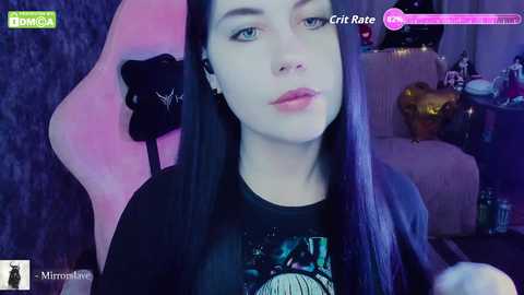 A video of a young woman with pale skin, long black hair, and blue eyes, wearing a black T-shirt with a cosmic graphic, sitting in a pink gaming chair with a black headrest.