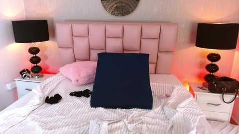 Video of a neatly made bed with a navy blue cushion, pink pillows, white striped sheets, and a white nightstand with black lamps and red lighting.