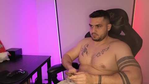 Video of a muscular, shirtless man with dark hair, tattoos, and a beard, seated in a black gaming chair, with purple and pink lighting in the background.