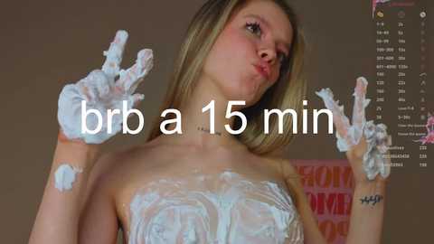 Media: Video of a blonde woman covered in white shaving cream, making peace signs, with \"brr a 15 min\" overlayed. Background is blurred with a \"HOMEWORK\" sign.