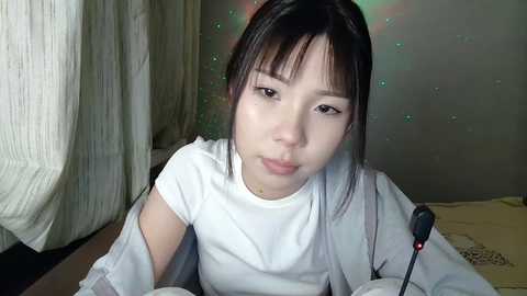 A video of an East Asian woman with straight, black hair, wearing a white top, lying on a bed, looking into the camera. The background features light-colored curtains and a soft, glowing light effect.