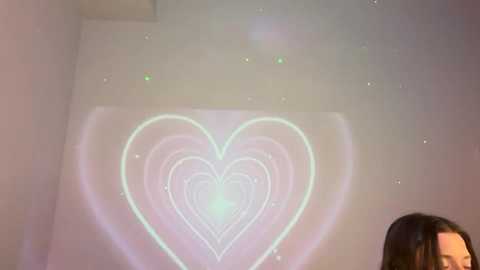 Video of a woman with dark hair, standing in a dimly lit room with a glowing heart-shaped light on the wall, casting a soft, warm glow.