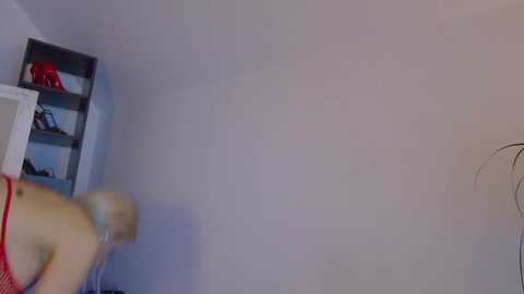 A video of a pale-skinned, blonde woman in a red top, blurry due to movement, captured from the side. She stands near a white shelf with a red bag and a white plant. The room has a minimalist, clean look with plain walls.