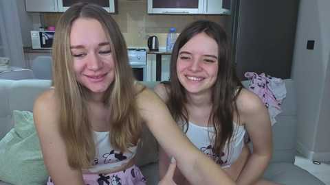 Video of two young, smiling Caucasian women in matching pink Minnie Mouse tank tops, sitting on a light grey couch in a modern, minimalistic kitchen.