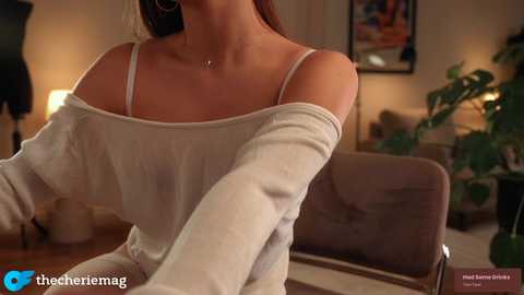 Video of a fair-skinned woman with light brown hair, wearing a white off-shoulder top and a delicate necklace, in a cozy, warmly lit living room with potted plants and framed artwork.