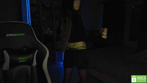 Video of a dimly lit room featuring a gamer with long hair wearing a black hoodie and shorts, holding a controller. A gaming chair with green accents and the logo \"MMAN\" is visible in the foreground.