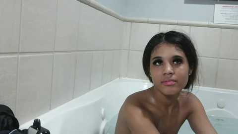 Video of a young, topless Latina woman with dark hair, sitting in a white bathtub, looking contemplative. Background features tiled walls and a partially visible sign.