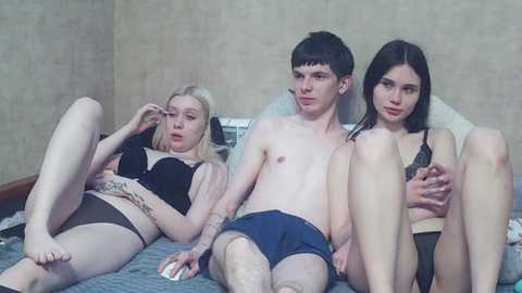 Video of a young, pale-skinned man with short dark hair and tattoos, wearing blue boxer shorts, sitting between a blonde woman in a black bra and panties, and a brunette woman in a black bra, all on a bed.