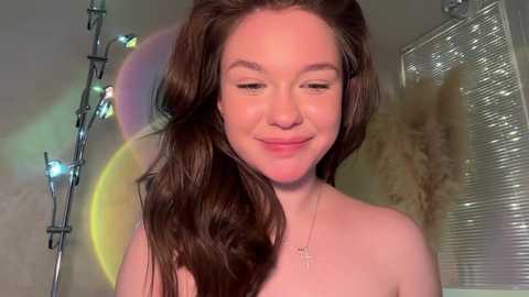 A video of a fair-skinned young woman with long, wavy brown hair, wearing a cross necklace, smiling softly, with a faint rainbow lens flare in the background.