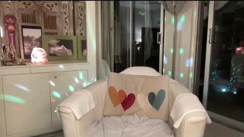 Video of a cozy living room at night, featuring a white leather armchair with a colorful heart pillow, surrounded by decorative items, framed photos, and a lit-up tree, creating a warm, festive atmosphere.