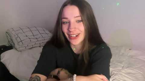 Video of a smiling young woman with long dark hair, wearing a black t-shirt, sitting on a bed with white sheets and a patterned pillow.