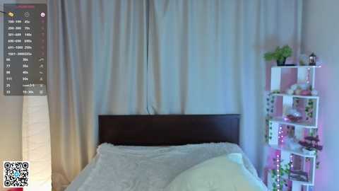 A video of a cozy bedroom with a dark wooden headboard, light gray bedding, and beige curtains. A white shelving unit on the right holds small plants and decorative items. A QR code is visible in the bottom left corner.