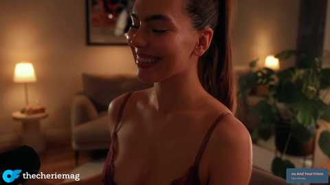Video of a smiling woman with dark hair tied back, wearing a maroon lace bralette, in a cozy, dimly-lit living room with a potted plant and two lamps.