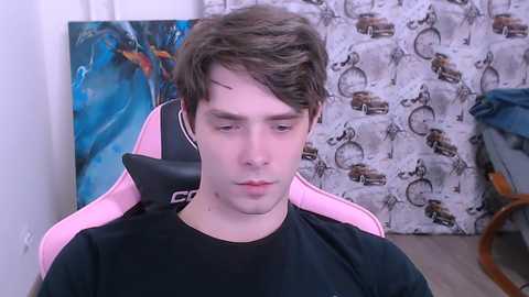 Video of a young man with short brown hair, wearing a black shirt, seated in a pink gaming chair. Background features a blue and orange abstract painting and a white curtain with vintage car patterns.