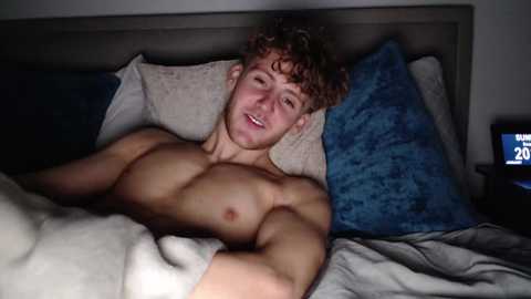 Media: Video of a shirtless, muscular young man with curly brown hair, lying on a bed with blue and white pillows, smiling.