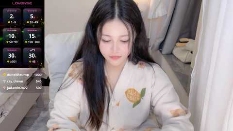 Media: Video of a young East Asian woman with long black hair, wearing a floral robe, seated indoors. Background includes a bed and a white curtain. A digital screen displays live stream metrics.