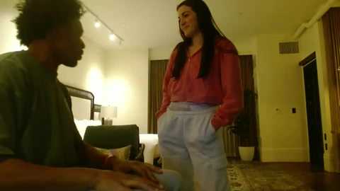 Video of a young woman with long dark hair, wearing a pink blouse and high-waisted light blue pants, standing in a modern, warmly lit hotel room.