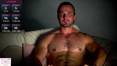 Video of a shirtless, muscular white man with short dark hair, lying on a bed with a grey wall background, displaying fitness stats on a virtual screen.