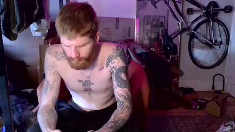 Video of a shirtless, tattooed, red-haired man with a beard, sitting on a wooden bench, surrounded by cluttered, dimly-lit room with bicycles and boxes.