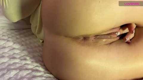 A close-up video of a nude, light-skinned woman lying on a bed, showing her shaved vulva with her fingers gently parting the labia. The background is a soft, patterned bedspread.