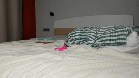 A video of a messy bed in a modern bedroom with a white headboard, green and white striped bedding, and a pink toy on the white comforter.