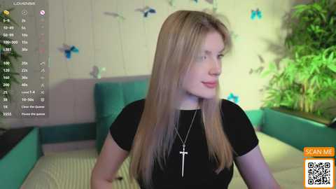 Video of a fair-skinned woman with long blonde hair, wearing a black top and necklace, sitting in a room with a green couch and plant in the background.