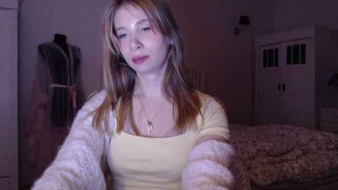 Video of a young Caucasian woman with light skin, straight brown hair, and a medium build, wearing a yellow top and white cardigan, sitting on a bed with a floral blanket in a dimly lit bedroom.